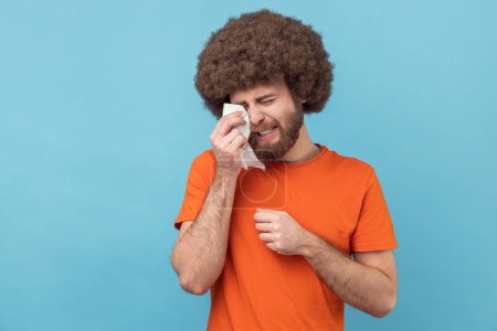 Photo for Portrait of depressed man with Afro hairstyle wearing orange T-shirt holding head down, hiding his face in hand and crying, feeling desperate. Indoor studio shot isolated on blue background. - Royalty Free Image