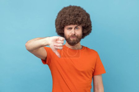 Photo for Portrait of displeased man with Afro hairstyle wearing orange T-shirt showing thumbs down dislike gesture, symbol of disagree, giving feedback. Indoor studio shot isolated on blue background. - Royalty Free Image