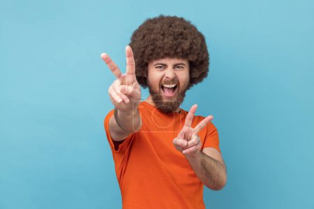 Téléchargez les photos : Portrait d'un bel homme excité et joyeux avec une coiffure afro portant un T-shirt orange montrant un signe de paix, un geste en v et criant joyeusement. Studio intérieur tourné isolé sur fond bleu. - en image libre de droit