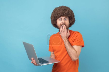 Photo for Portrait of man with Afro hairstyle wearing orange T-shirt working on notebook, sees something astonishing, covering mouth with palm. Indoor studio shot isolated on blue background. - Royalty Free Image