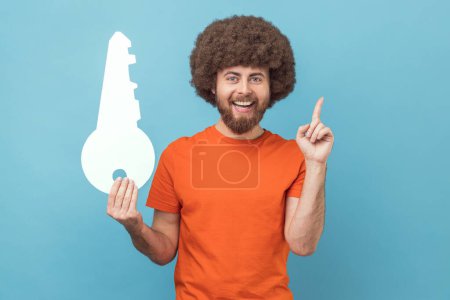 Téléchargez les photos : Portrait d'homme souriant à la coiffure afro portant un T-shirt orange tenant une grande clé en papier et pointant du doigt, ayant l'idée de louer un nouvel appartement. Studio intérieur tourné isolé sur fond bleu. - en image libre de droit