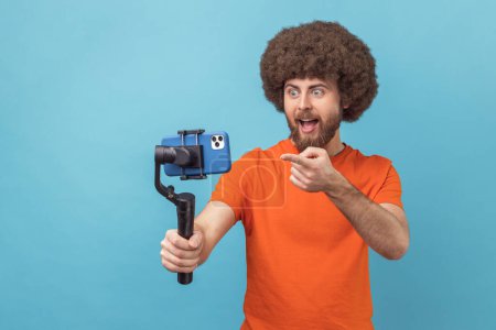Photo for Portrait of astonished man with Afro hairstyle wearing orange T-shirt using stedicam and phone for livestream, being amazed, pointing at phone camera. Indoor studio shot isolated on blue background. - Royalty Free Image