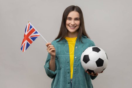 Téléchargez les photos : Heureuse femme positive tenant dans les mains le drapeau du Royaume-Uni et le ballon de football, soutenant l'équipe préférée au championnat, portant une veste de style décontracté. Studio intérieur tourné isolé sur fond gris. - en image libre de droit