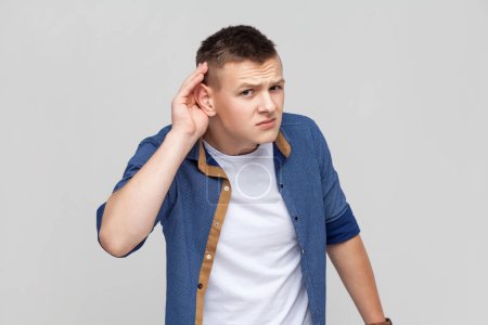 Téléchargez les photos : Portrait d'un adolescent portant une chemise bleue tenant la main près de l'oreille et écoutant attentivement, ayant des problèmes d'audition, surdité dans la communication. Studio intérieur tourné isolé sur fond gris. - en image libre de droit