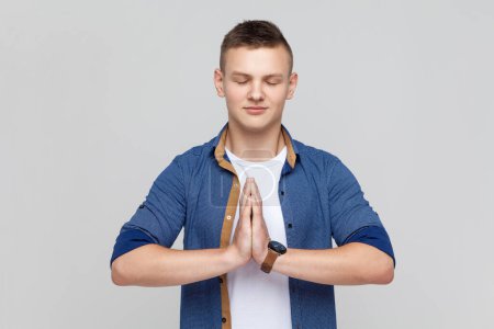 Photo for Portrait of calm attractive smiling teenager boy wearing blue shirt standing in yoga pose and try to relaxing, keeps palms together. Indoor studio shot isolated on gray background. - Royalty Free Image