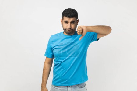 Photo for Portrait of unshaven man wearing blue T- shirt standing assess project, shows sign of dislike, looks with negative expression and disapproval. Indoor studio shot isolated on gray background. - Royalty Free Image