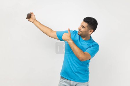 Photo for Portrait of joyful satisfied delighted man blogger in blue T- shirt holding mobile phone, has video call or livestream, showing thumb up, like gesture. Indoor studio shot isolated on gray background. - Royalty Free Image