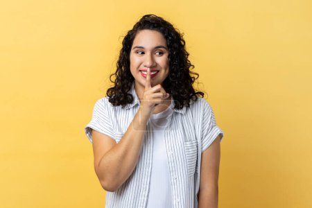 Photo for Portrait of attractive woman with dark wavy hair standing, showing hush sign and looking away with toothy smile, sharing secret. Indoor studio shot isolated on yellow background. - Royalty Free Image