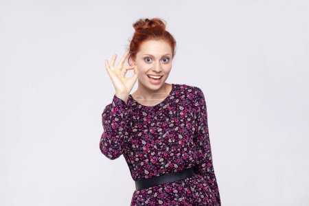 Téléchargez les photos : Portrait d'une jeune femme rousse joyeuse et joyeuse portant une robe montrant un panneau ok, regardant la caméra avec un sourire de dents. Studio intérieur tourné isolé sur fond gris. - en image libre de droit