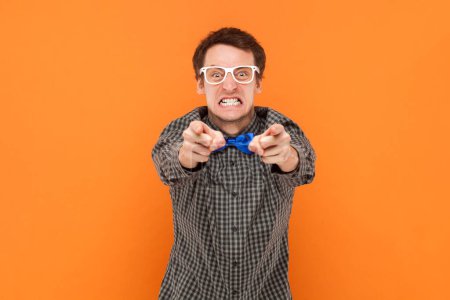 Téléchargez les photos : Portrait d'un homme fou agressif en colère pointant vers vous, regardant la caméra avec des dents serrées, portant une chemise avec un noeud papillon bleu et des lunettes blanches. Studio intérieur tourné isolé sur fond orange - en image libre de droit