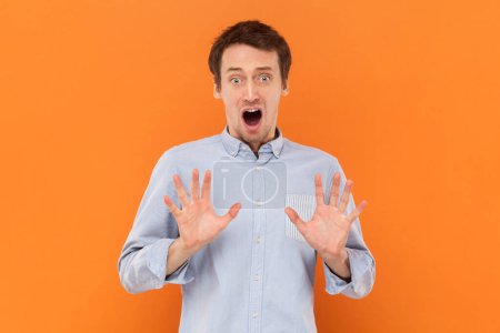 Photo for Portrait of scared frighten young adult man standing with raised arms, showing stop gesture, sees something scary, wearing light blue shirt. Indoor studio shot isolated on orange background. - Royalty Free Image