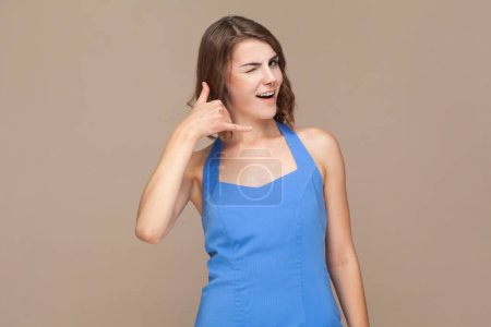 Photo for Call me back sign. Portrait of woman with wavy hair makes telephone gesture, asks to call her, expresses positive emotions, wearing blue dress. Indoor studio shot isolated on light brown background. - Royalty Free Image