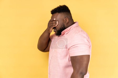 Photo for Side view of man wearing pink shirt closing eyes with hand, dont want to see that, ignoring problems, hiding from stressful situations. Indoor studio shot isolated on yellow background. - Royalty Free Image