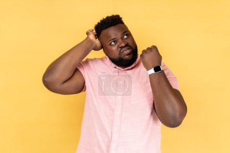 Photo for Portrait of puzzled confused man wearing pink shirt showing his smart watch and scratching his head, looking away with with thoughtful expression. Indoor studio shot isolated on yellow background. - Royalty Free Image