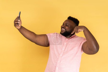 Téléchargez les photos : Portrait d'un bel homme positif portant une chemise rose prenant selfie ou ayant livestream, pointant du doigt vers le bas, abonnez-vous à son vlog. Studio intérieur tourné isolé sur fond jaune. - en image libre de droit