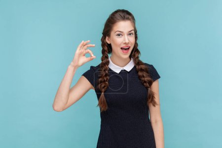 Photo for Portrait of beautiful woman with braids making okay gesture, demonstrates agreement, likes idea, smiles happily, wearing black dress. woman Indoor studio shot isolated on blue background. - Royalty Free Image