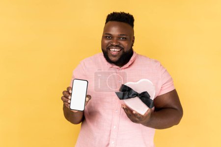 Photo for Portrait of joyful cheerful bearded man wearing pink shirt holding heart shaped present box and showing mobile phone with empty display. Indoor studio shot isolated on yellow background. - Royalty Free Image