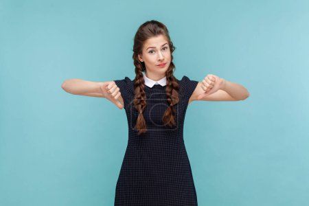 Photo for Portrait of displeased naughty woman with braids standing and showing thumb down, dislike bad service, not recommend, wearing black dress. woman Indoor studio shot isolated on blue background. - Royalty Free Image