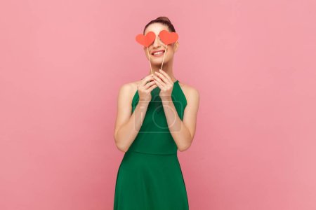 Photo for Portrait of charming unknown woman with bun hairstyle standing and covering her eyes with little red hearts on stick, smiling, wearing green dress. Indoor studio shot isolated on pink background. - Royalty Free Image