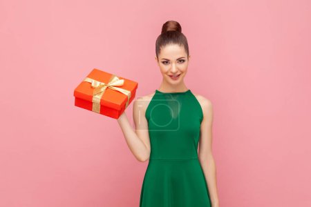 Photo for Portrait of attractive positive smiling woman standing with red present box, looking at camera, celebrating holiday, wearing green dress. Indoor studio shot isolated on pink background. - Royalty Free Image