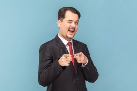 Photo for Portrait of flirting positive optimistic man with mustache standing pointing to camera, choosing you, wearing black suit with red tie. Indoor studio shot isolated on light blue background. - Royalty Free Image
