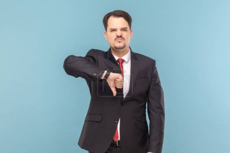 Photo for Displeased sad showing man with mustache standing showing dislike gesture, grimacing, feels dissatisfied, wearing black suit with red tie. Indoor studio shot isolated on light blue background. - Royalty Free Image