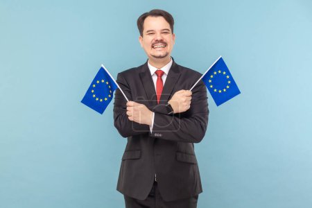 Téléchargez les photos : Portrait d'un homme portant des drapeaux européens, symbole à la fois de l'Union européenne et de l'identité et de l'unité de l'Europe, vêtu d'un costume noir avec cravate rouge. Studio intérieur tourné isolé sur fond bleu clair. - en image libre de droit