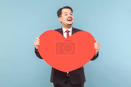 Photo for Portrait of satisfied joyful man with mustache standing holding big red card, expressing love and gentle, wearing black suit with red tie. Indoor studio shot isolated on light blue background. - Royalty Free Image