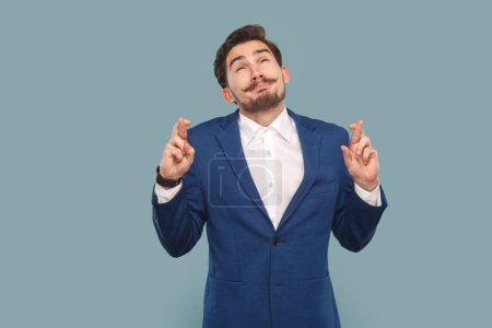 Photo for Portrait of hopeful handsome man with mustache standing with crossed fingers, hopes for better and good luck, wearing white shirt and jacket. Indoor studio shot isolated on light blue background. - Royalty Free Image