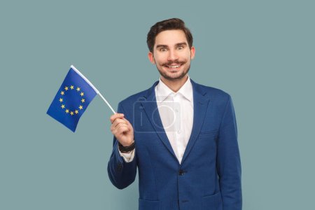 Téléchargez les photos : Portrait de sourire heureux bel homme avec moustache debout agitant drapeau de l'Union européenne, exprimant le bonheur, portant chemise blanche et veste. Studio intérieur tourné isolé sur fond bleu clair. - en image libre de droit
