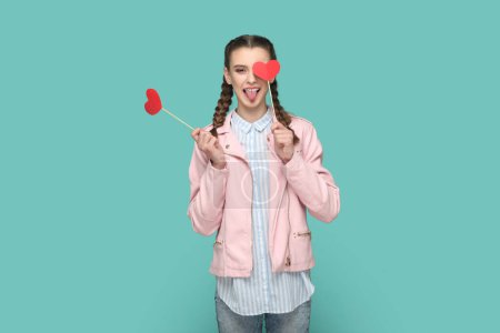 Téléchargez les photos : Portrait d'adolescente romantique positive avec des tresses portant une veste rose debout couvrant l'œil avec un cœur rouge sur le bâton, montrant la langue. Studio intérieur tourné isolé sur fond vert. - en image libre de droit