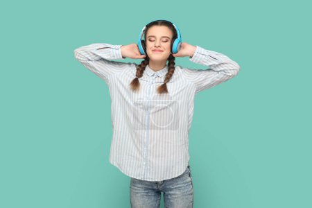Téléchargez les photos : Portrait d'une adolescente avec des tresses portant une chemise rayée debout écoutant de la musique préférée avec des écouteurs, les yeux fermés et souriant. Studio intérieur tourné isolé sur fond vert. - en image libre de droit
