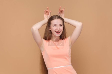 Photo for Portrait of funny smiling joyful woman with long hair standing with raised arms, showing horns, looking at camera, wearing elegant dress. Indoor studio shot isolated on brown background. - Royalty Free Image