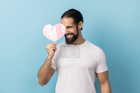 Téléchargez les photos : Portrait d'homme barbu portant un T-shirt blanc tenant et couvrant l'œil avec du papier rose coeur sur bâton et souriant à la caméra, partageant l'amour. Studio intérieur tourné isolé sur fond bleu. - en image libre de droit