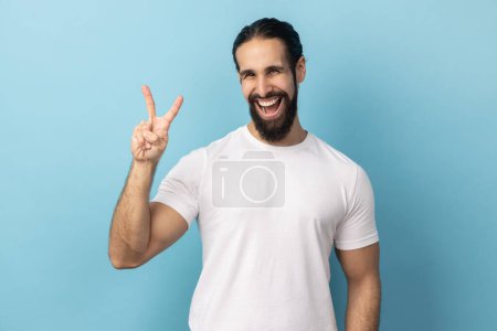 Photo for Portrait of positive man with beard wearing white T-shirt looking at camera and gesturing victory sign at camera, success or achievement. Indoor studio shot isolated on blue background. - Royalty Free Image