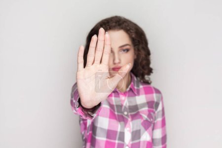 Photo for Woman with curly hair showing stop sign, prohibition symbol, keeps palm forward to camera, looks with strict expression, wearing pink checkered shirt. Indoor studio shot isolated on gray background. - Royalty Free Image