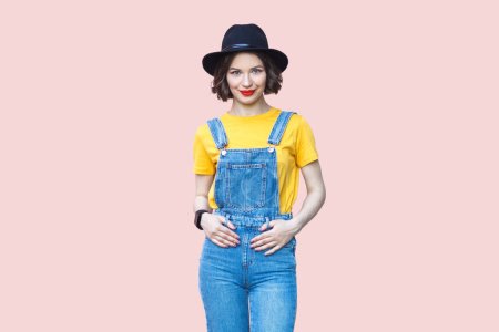 Téléchargez les photos : Portrait d'une joyeuse femme hipster souriante en salopette en denim bleu, T-shirt jaune et chapeau noir regardant la caméra avec une expression heureuse. Studio intérieur isolé sur fond rose clair. - en image libre de droit