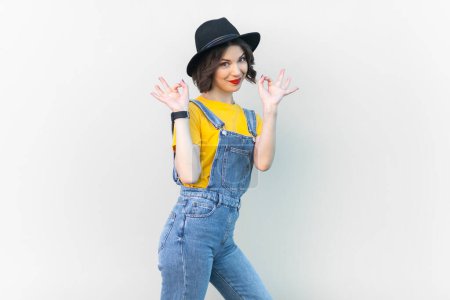 Téléchargez les photos : Portrait de femme optimiste heureuse hipster en combinaison en denim bleu, T-shirt jaune et chapeau noir, levé les bras, montrant un signe correct. Studio intérieur tourné isolé sur fond gris. - en image libre de droit