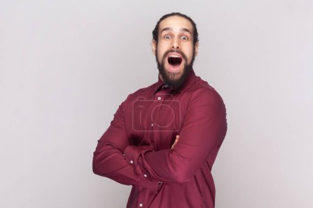 Photo for Portrait of amazed surprised man with dark hair and beard in red shirt standing with crossed arms, looking at camera with big eyes. Indoor studio shot isolated on gray background. - Royalty Free Image
