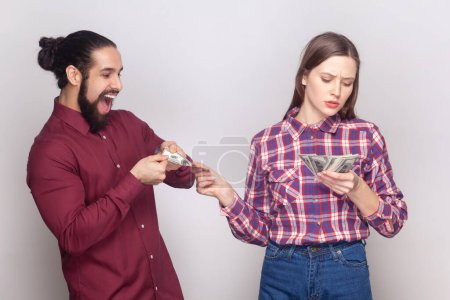 Photo for Portrait of serious rich confident woman and man gigolo standing together, wife giving money to her husband with indifferent face. Indoor studio shot isolated on gray background. - Royalty Free Image