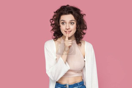 Photo for Portrait of positive smiling woman with curly hairstyle wearing casual style outfit showing shh gesture, asking to keep secret. Indoor studio shot isolated on pink background. - Royalty Free Image