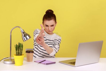 Photo for Angry woman office worker sitting at workplace with laptop threatening and pointing finger gun to camera, shooting with aggressive expression. Indoor studio studio shot isolated on yellow background. - Royalty Free Image