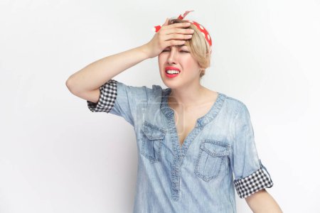 Photo for Portrait of sorrowful depressed blonde young woman wearing blue denim shirt and red headband standing making showing facepalm gesture. Indoor studio shot isolated on gray background. - Royalty Free Image