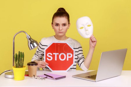 Photo for Portrait of serious dark haired woman holding white mask with unknown face and red traffic sign while sitting on workplace with laptop. Indoor studio studio shot isolated on yellow background. - Royalty Free Image
