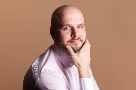 Photo for Side view portrait of cute positive smiling bald bearded man sitting holding chin, looking at camera with positive expression, wearing light pink shirt. Indoor studio shot isolated on brown background - Royalty Free Image
