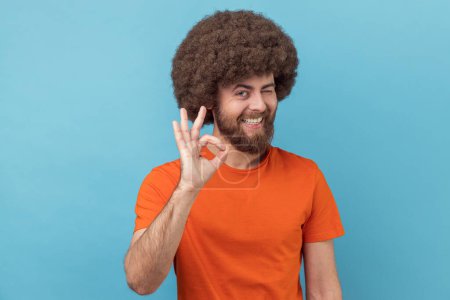 Photo for Portrait of positive man with Afro hairstyle wearing orange T-shirt standing and looking at camera with Ok sign, winking and toothy smile. Indoor studio shot isolated on blue background. - Royalty Free Image
