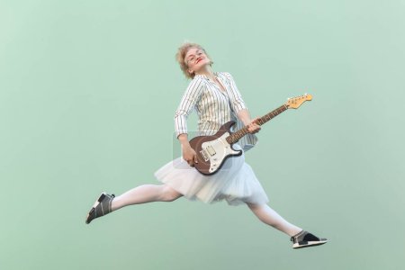 Photo for Portrait of young adult happy blonde woman in white shirt, skirt, and striped blouse with eyeglasses holding electric guitar, smiling and jumping. Indoor studio shot isolated on light green background - Royalty Free Image