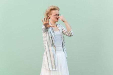 Téléchargez les photos : Portrait d'une femme blonde dégoûtée et confuse portant une chemise rayée et une jupe pincant le nez, montrant un geste d'arrêt. Studio intérieur tourné isolé sur fond vert clair. - en image libre de droit