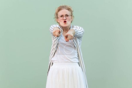 Photo for Portrait of angry shocked young adult blonde woman wearing striped shirt and skirt, pointing at you, choosing you, screaming. Indoor studio shot isolated on light green background. - Royalty Free Image