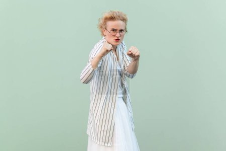 Téléchargez les photos : Portrait d'une femme blonde agressive et agressive en colère portant une chemise et une jupe rayées, serrant les poings, prête à attaquer. Studio intérieur tourné isolé sur fond vert clair. - en image libre de droit